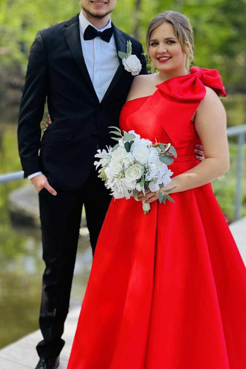 Girl standing by her prom date with a bright red dress and a bow on the shoulder.