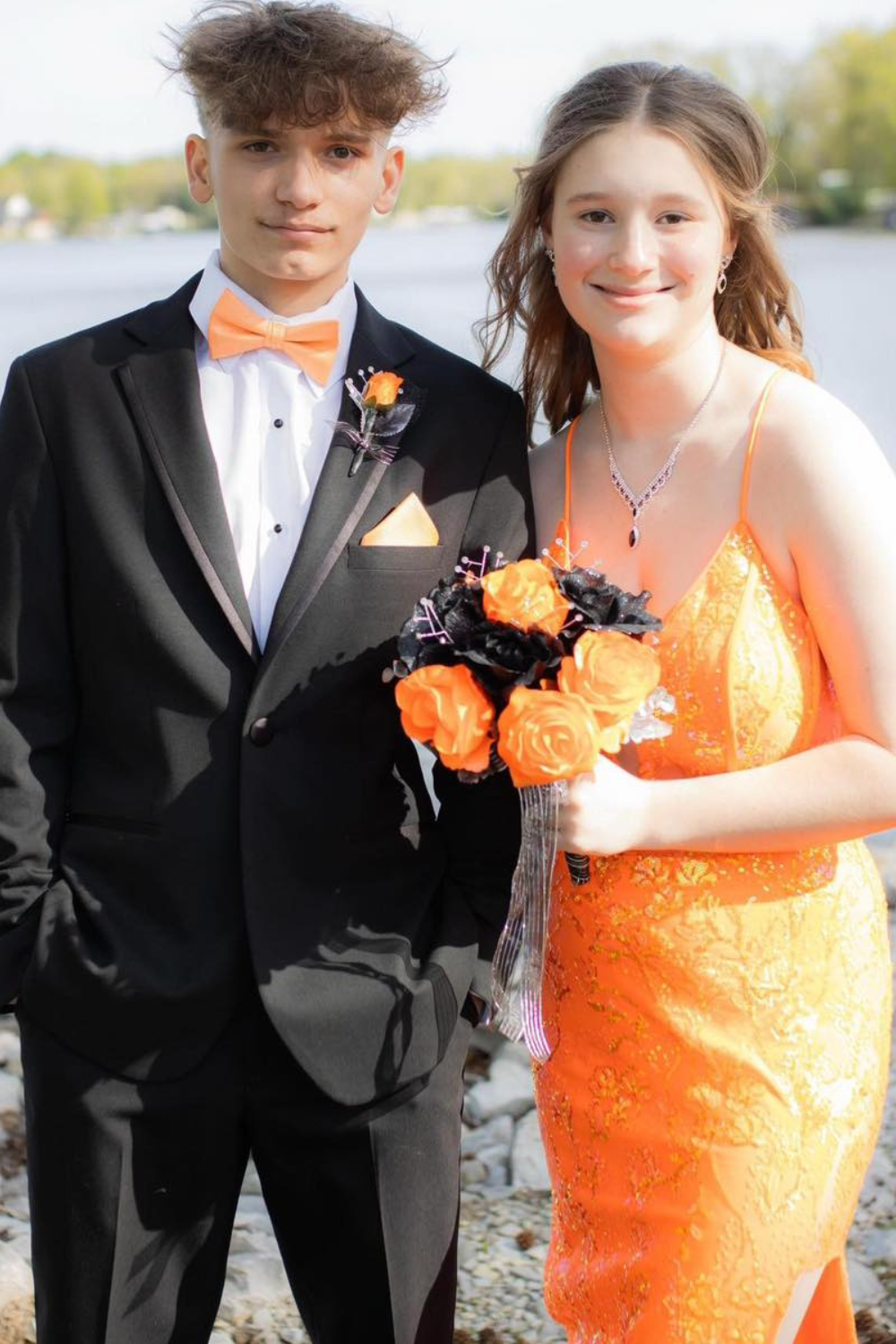 A prom couple standing in front of a lake with matching orange dress and bow tie.