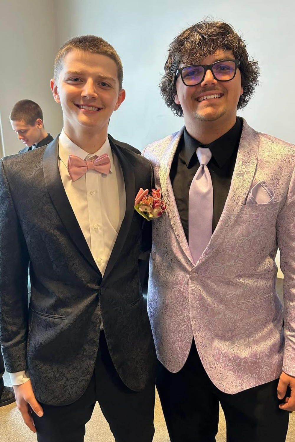 Two boys standing beside each other with one wearing a grey suit with white dress shirt and the other wearing a silver coat and tie and black pants.