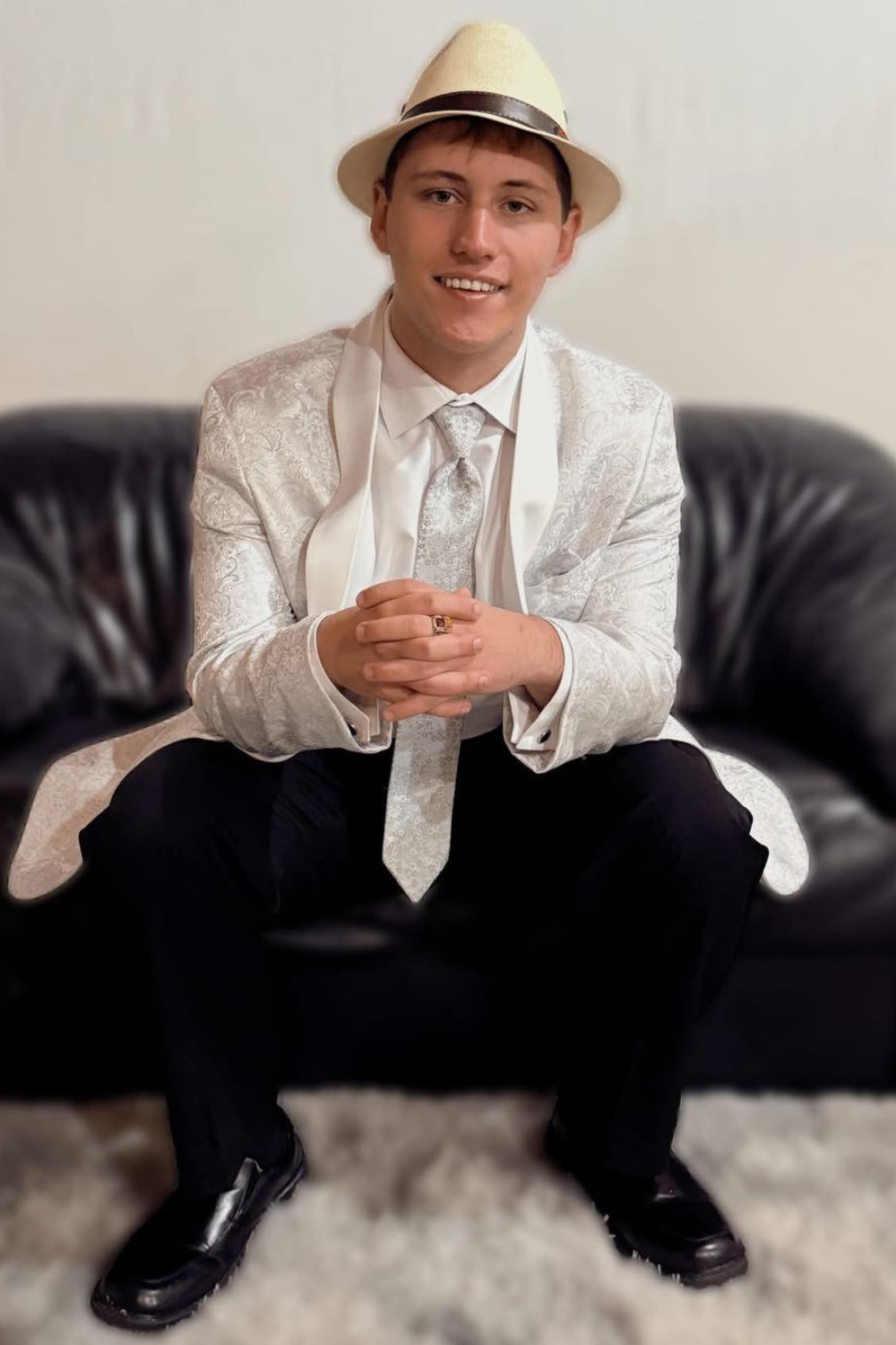 A boy sitting on a cound in a tux with a silver sports coat and beige fedora.