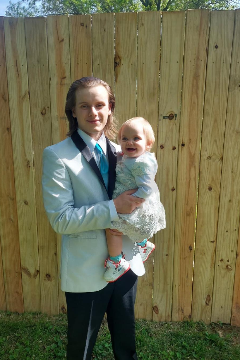 Boy with a white tuxedo sportcoat and black pants holding an infant.