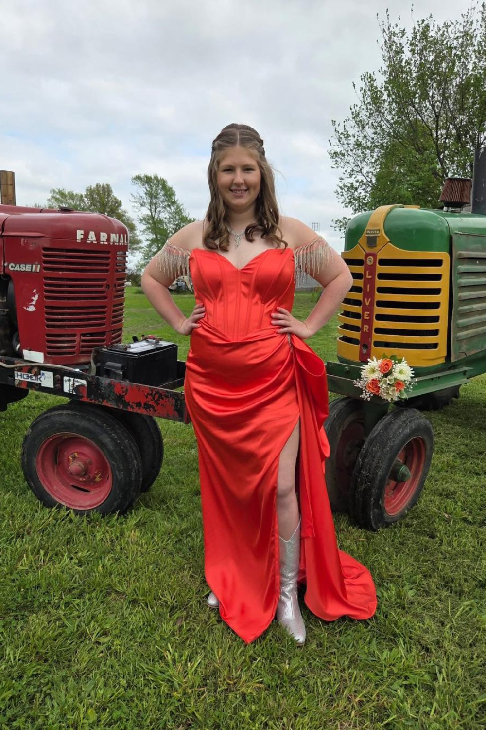 A girl with a red dress with sequins and a slit in the side standing between two tractors.