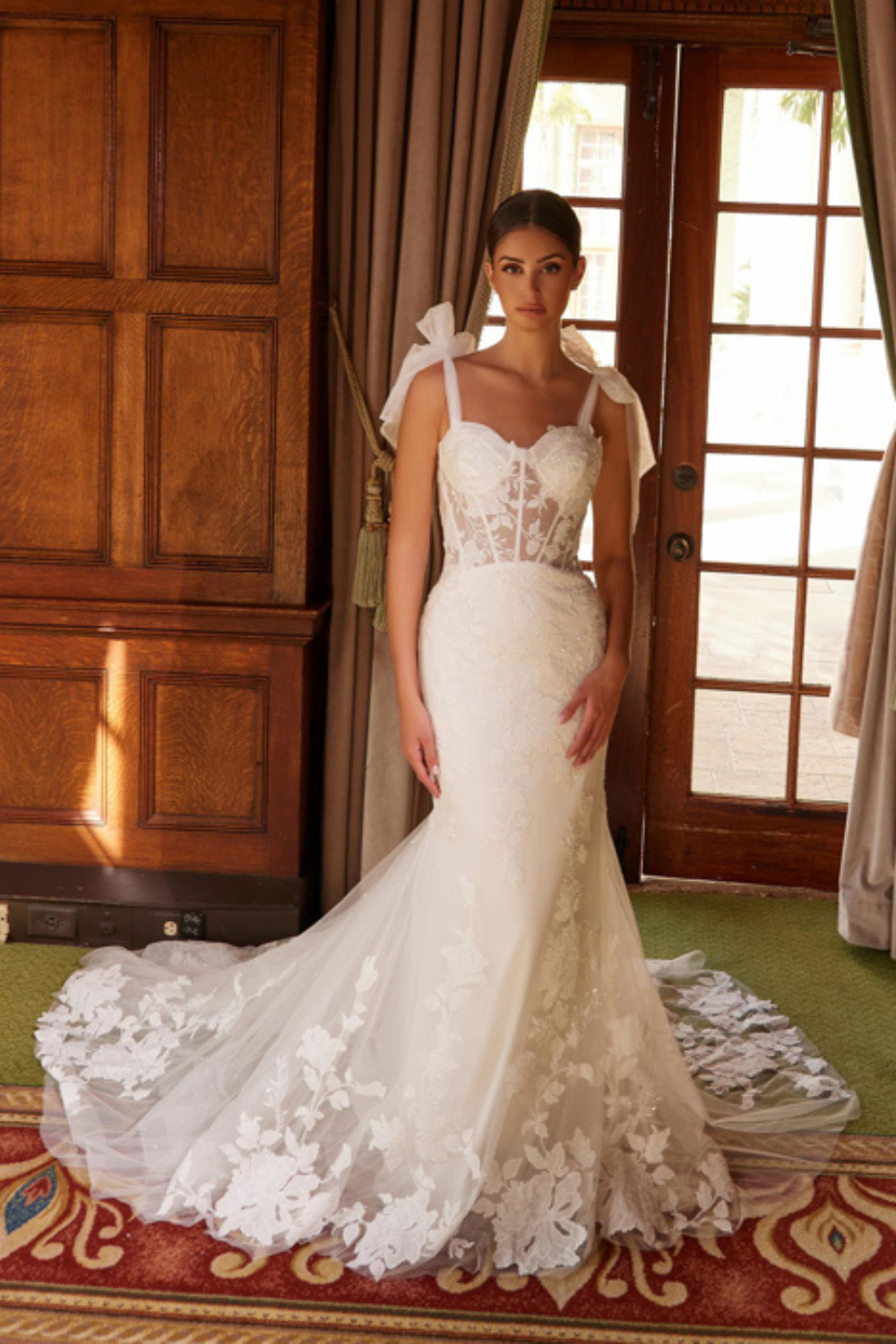 Woman standing in front of french doors in a white lace wedding dress with bows tied on the shoulders. 