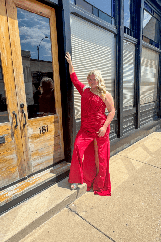 Home 3 Black Tie Affair model with blonde hair in red one sleeved gown with a slit in the leg.